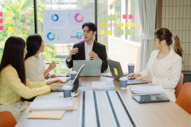 Asyalı iş adamlarından oluşan bir grup konferans odasında meslektaşlarına veri sunuyor. Takım elbiseli işadamları, şirket verilerini sunmak için masadaki grafik dokümanlarına kalem tutuyorlar..