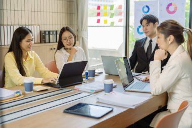 Asyalı iş adamlarından oluşan bir grup konferans odasında meslektaşlarına veri sunuyor. Takım elbiseli işadamları, şirket verilerini sunmak için masadaki grafik dokümanlarına kalem tutuyorlar..