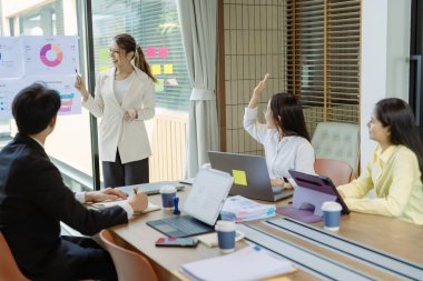 Asyalı iş adamlarından oluşan bir grup konferans odasında meslektaşlarına veri sunuyor. Takım elbiseli işadamları, şirket verilerini sunmak için masadaki grafik dokümanlarına kalem tutuyorlar..