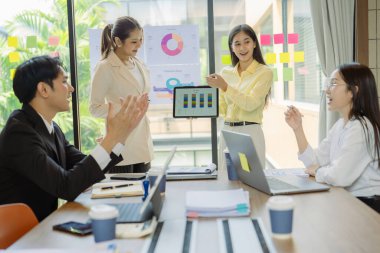 Asyalı iş adamlarından oluşan bir grup konferans odasında meslektaşlarına veri sunuyor. Takım elbiseli işadamları, şirket verilerini sunmak için masadaki grafik dokümanlarına kalem tutuyorlar..