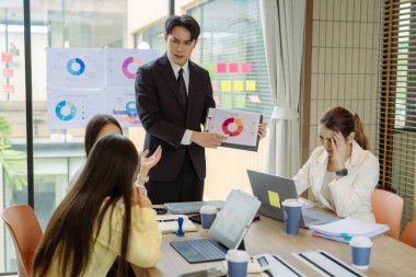 Asyalı iş adamlarından oluşan bir grup konferans odasında meslektaşlarına veri sunuyor. Takım elbiseli işadamları, şirket verilerini sunmak için masadaki grafik dokümanlarına kalem tutuyorlar..