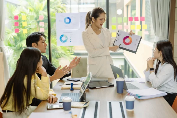 Asyalı iş adamlarından oluşan bir grup konferans odasında meslektaşlarına veri sunuyor. Takım elbiseli işadamları, şirket verilerini sunmak için masadaki grafik dokümanlarına kalem tutuyorlar..