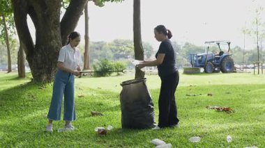 İki Asyalı kadın parkta çöp torbalarıyla plastik şişe topluyor ve park alanını temizlemenin keyfini çıkarıyorlar..