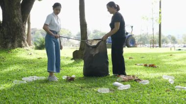 İki Asyalı kadın parkta çöp torbalarıyla plastik şişe topluyor ve park alanını temizlemenin keyfini çıkarıyorlar..
