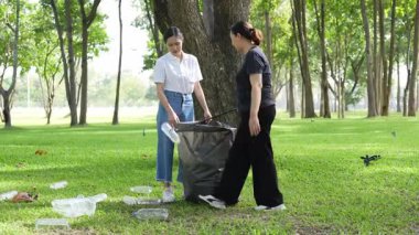 İki Asyalı kadın parkta çöp torbalarıyla plastik şişe topluyor ve park alanını temizlemenin keyfini çıkarıyorlar..