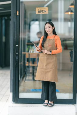 Önlüklü genç Asyalı kadın kahve dükkanında dijital tablet tutuyor. Barista, garson gülümseyip kameraya bakıyor..