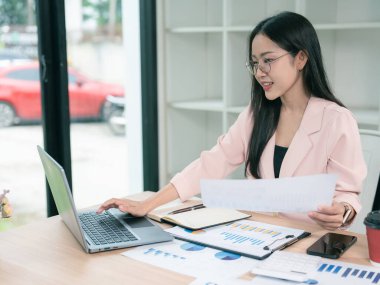 Bilgisayarla ve finansal belgelerle çalışan Asyalı güzel bir iş kadını ofiste dizüstü bilgisayarla analiz hesaplıyor..