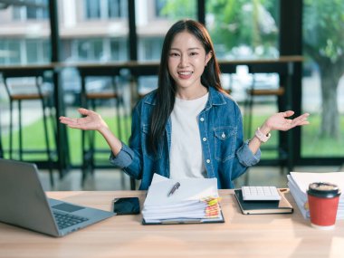 Asyalı bir kadın ofiste dizüstü bilgisayarla çalışıyor, elinde kalem tutuyor ve finansal belgeler okuyor, gülümsüyor ve bilgisayarla ofiste çalışıyor..