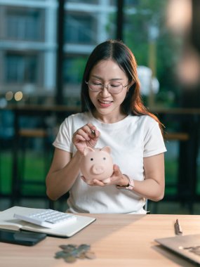 Asyalı kadın para biriktirmek için domuz kumbarasına para koyuyor, gelecekteki kullanım için finansal planlar yapıyor..