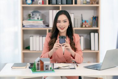Young Asian woman sitting in office working on laptop, holding keys and house model in front for real estate concept.