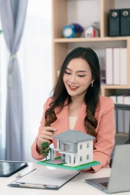 Young Asian woman sitting in office working on laptop, holding keys and house model in front for real estate concept.