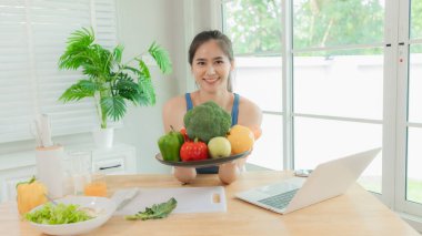 Sağlıklı genç Asyalı kadın dizüstü bilgisayar kullanıyor ve mutfakta havuç tutuyor. Bilgisayardan arıyor ve sağlıklı beslenme alışkanlıklarını öğreniyor..