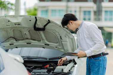 Genç Asyalı bir adam, bir kır yolunun kenarına park etmiş bozuk bir arabanın motorunu kontrol ediyor. Genç adam yolda bozuk bir arabanın kaputunu açıyor..