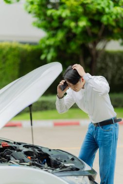 Genç Asyalı bir adam, bir kır yolunun kenarına park etmiş bozuk bir arabanın motorunu kontrol ediyor. Genç adam yolda bozuk bir arabanın kaputunu açıyor..