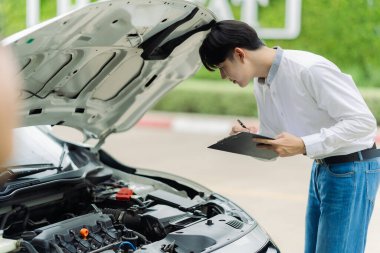 Genç Asyalı bir adam, bir kır yolunun kenarına park etmiş bozuk bir arabanın motorunu kontrol ediyor. Genç adam yolda bozuk bir arabanın kaputunu açıyor..