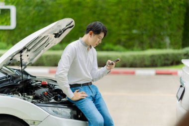 Genç Asyalı bir adam, bir kır yolunun kenarına park etmiş bozuk bir arabanın motorunu kontrol ediyor. Genç adam yolda bozuk bir arabanın kaputunu açıyor..