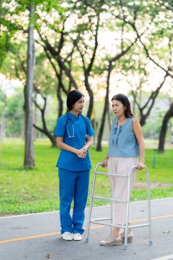 Kadın bir doktor, yaşlı bir Asyalı kadına yeşil doğada cesaret ve rahatlık vererek bastonla yürümeyi öğrenmesine yardım eder..