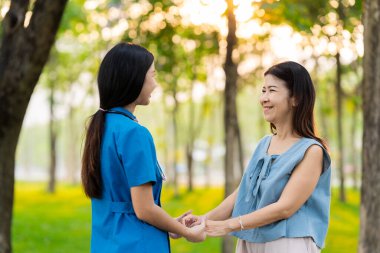 Asyalı hemşire ve yaşlı kadın hasta parkta el ele tutuşuyorlar. Emeklilikte yaşlılara bakım kavramı. Sağlık Hizmetleri Merkezi.