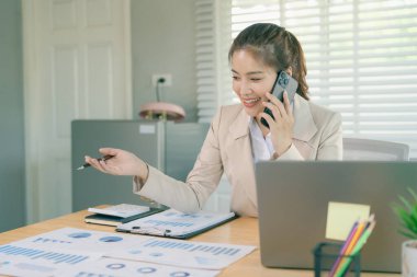 Mutlu bir iş kadını akıllı telefonunu ve laptopunu kullanıyor ve bir şirket ofisinde bilgisayar teknolojisi kullanarak finansal belgelerle uğraşıyor..