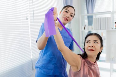 Bir Asyalı hemşire, fizik tedavi sırasında direnç bantları kullanarak hastaya egzersiz yaptırırken yardımcı olur. Hemşire de hastanın odasında yardımcı olur..