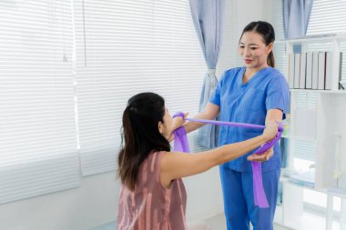 Bir Asyalı hemşire, fizik tedavi sırasında direnç bantları kullanarak hastaya egzersiz yaptırırken yardımcı olur. Hemşire de hastanın odasında yardımcı olur..