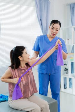 Bir Asyalı hemşire, fizik tedavi sırasında direnç bantları kullanarak hastaya egzersiz yaptırırken yardımcı olur. Hemşire de hastanın odasında yardımcı olur..