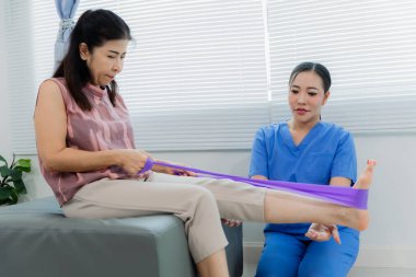 Bir Asyalı hemşire, fizik tedavi sırasında direnç bantları kullanarak hastaya egzersiz yaptırırken yardımcı olur. Hemşire de hastanın odasında yardımcı olur..