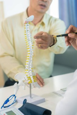 Asyalı bir erkek doktor yaşlı bir hastaya omurilik modeli tutarak ve hastanın ağrısının nedenini açıklayarak omurilik röntgeni görüntülerini açıklıyor..