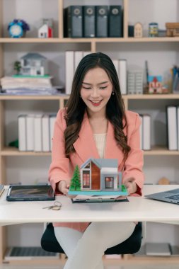 Asyalı bir kadın elinde model bir ev tutuyor ve önünde model bir ev olan bir dizüstü bilgisayarla çalışıyor. Gayrimenkul, iş ve finans kavramları için uygun.