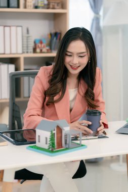 Asyalı bir kadın elinde model bir ev tutuyor ve önünde model bir ev olan bir dizüstü bilgisayarla çalışıyor. Gayrimenkul, iş ve finans kavramları için uygun.