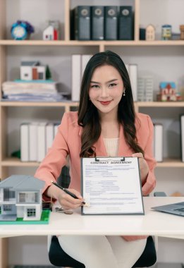 Asyalı bir kadın elinde model bir ev tutuyor ve önünde model bir ev olan bir dizüstü bilgisayarla çalışıyor. Gayrimenkul, iş ve finans kavramları için uygun.