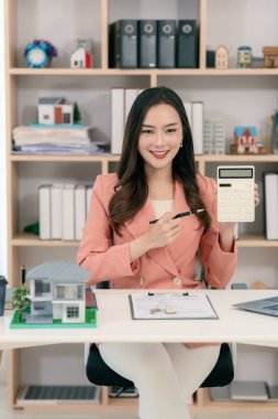 Asyalı bir kadın elinde model bir ev tutuyor ve önünde model bir ev olan bir dizüstü bilgisayarla çalışıyor. Gayrimenkul, iş ve finans kavramları için uygun.
