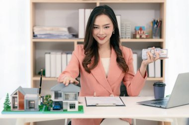 Asyalı bir kadın elinde model bir ev tutuyor ve önünde model bir ev olan bir dizüstü bilgisayarla çalışıyor. Gayrimenkul, iş ve finans kavramları için uygun.