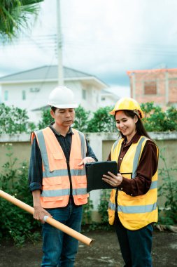 Asyalı erkek ve kadın mühendisler ve mimarlar inşaat alanında tartışıyorlar; güvenlik yeleği giyen profesyonel meslektaşlar sanayi tipi konut projesinde işbirliği yapıyorlar.