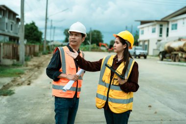 Asyalı erkek ve kadın mühendisler ve mimarlar inşaat alanında tartışıyorlar; güvenlik yeleği giyen profesyonel meslektaşlar sanayi tipi konut projesinde işbirliği yapıyorlar.