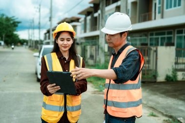 Asyalı erkek ve kadın mühendisler ve mimarlar inşaat alanında tartışıyorlar; güvenlik yeleği giyen profesyonel meslektaşlar sanayi tipi konut projesinde işbirliği yapıyorlar.