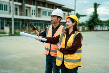 Asyalı erkek ve kadın mühendisler ve mimarlar inşaat alanında tartışıyorlar; güvenlik yeleği giyen profesyonel meslektaşlar sanayi tipi konut projesinde işbirliği yapıyorlar.