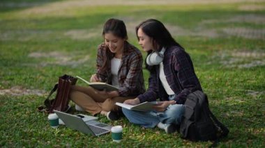 İki gülümseyen Asyalı kız öğrenci bir üniversite parkında dizüstü bilgisayarları, çalışmaları, teknolojileri ve insanlarıyla oturuyorlar..
