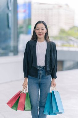 Güzel bir Asyalı kadın bir alışveriş merkezinde şık bir şekilde dolaşırken, alışveriş torbaları taşırken ve şık bir kıyafet giyerken akıllı telefonunun keyfini çıkarırken görülüyor..