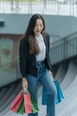 Güzel bir Asyalı kadın bir alışveriş merkezinde şık bir şekilde dolaşırken, alışveriş torbaları taşırken ve şık bir kıyafet giyerken akıllı telefonunun keyfini çıkarırken görülüyor..