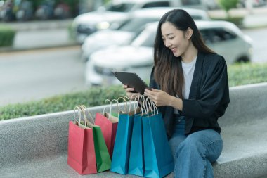 Güzel bir Asyalı kadın bir alışveriş merkezinde şık bir şekilde dolaşırken, alışveriş torbaları taşırken ve şık bir kıyafet giyerken akıllı telefonunun keyfini çıkarırken görülüyor..