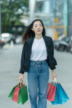 Güzel bir Asyalı kadın bir alışveriş merkezinde şık bir şekilde dolaşırken, alışveriş torbaları taşırken ve şık bir kıyafet giyerken akıllı telefonunun keyfini çıkarırken görülüyor..