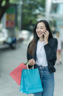 Güzel bir Asyalı kadın bir alışveriş merkezinde şık bir şekilde dolaşırken, alışveriş torbaları taşırken ve şık bir kıyafet giyerken akıllı telefonunun keyfini çıkarırken görülüyor..