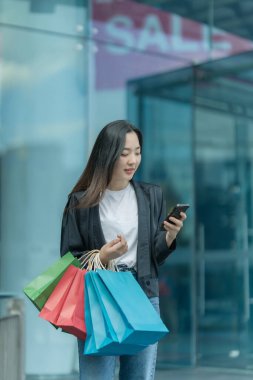 Güzel bir Asyalı kadın bir alışveriş merkezinde şık bir şekilde dolaşırken, alışveriş torbaları taşırken ve şık bir kıyafet giyerken akıllı telefonunun keyfini çıkarırken görülüyor..