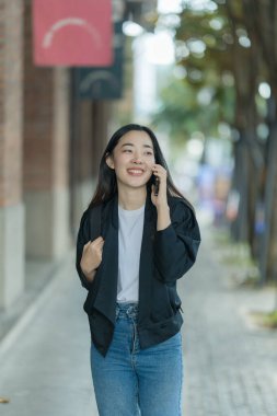 Güzel bir Asyalı kadın akıllı telefon ve sırt çantasıyla bir şehir caddesinde yürüyor, şehrin arka planında poz veriyor ve sosyal medya paylaşımı yaratıyor..