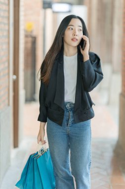 A beautiful Asian woman is seen strolling stylishly through a shopping mall, carrying shopping bags and enjoying her smartphone while wearing a trendy outfit.