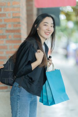 Güzel bir Asyalı kadın bir alışveriş merkezinde şık bir şekilde dolaşırken, alışveriş torbaları taşırken ve şık bir kıyafet giyerken akıllı telefonunun keyfini çıkarırken görülüyor..