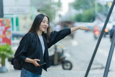Güzel bir Asyalı kadın akıllı telefon ve sırt çantasıyla bir şehir caddesinde yürüyor, şehrin arka planında poz veriyor ve sosyal medya paylaşımı yaratıyor..