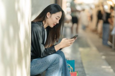 Güzel bir Asyalı kadın bir alışveriş merkezinde şık bir şekilde dolaşırken, alışveriş torbaları taşırken ve şık bir kıyafet giyerken akıllı telefonunun keyfini çıkarırken görülüyor..