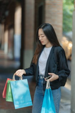 Güzel bir Asyalı kadın bir alışveriş merkezinde şık bir şekilde dolaşırken, alışveriş torbaları taşırken ve şık bir kıyafet giyerken akıllı telefonunun keyfini çıkarırken görülüyor..